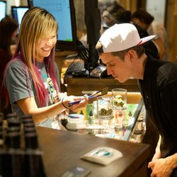 Lightshade Rec Dispensary - A smiling budtender with long blonde and pink hair assists a customer, holding cannabis with tweezers for him to smell inside...