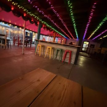 Twans Burger - The image shows an indoor bar area with a wooden bar counter and stools in yellow, green, and red. The ceiling is decorated with string lights in purple, green, yellow, and red, arranged in parallel lines. The space appears to be dimly lit, with some additional lighting along the side walls. There are tables and chairs visible in the background, and the overall setting suggests a casual, lively environment suitable for social gatherings. - Food Trucks Near Me - Denver, Colorado