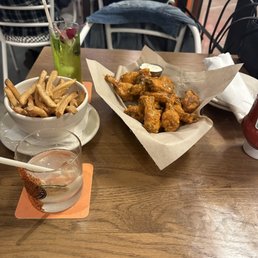 Mild buffalo wings, fries, 2 cocktails - The image shows a wooden table in a restaurant with a basket of French fries, a plate of fried chicken wings on parchment paper with a dipping sauce, a tall cocktail garnished with mint and fruit, and a glass of water with a straw placed on a coaster. In the background, there are other diners and chairs, indicating a casual dining environment.