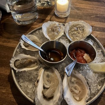 Prairie Fire - Oysters from the raw bar - A seafood platter featuring raw oysters on ice, accompanied by sauces, lemon wedges, and drinks, set on a wooden table with a candle and water glasses. - Pizza Near Me - Brookline, Massachusetts