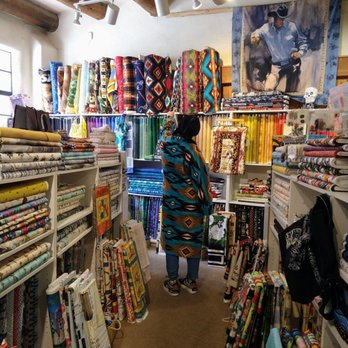 Taos Adobe Quilting - A woman stands in a fabric store aisle filled with colorful quilting fabrics and rolls, surrounded by shelves of fabric bolts and patterns. - Fabric Stores Near Me - Taos, New Mexico