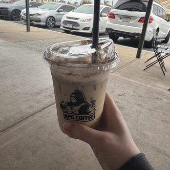 Ape Coffee - Tiramisu Latte - A person holds a plastic cup of iced coffee from Ape Coffee, with a parking lot and cars visible outside. - Cafes Near Me - Placentia, California