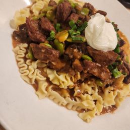 Watered stroganoff - A dish of beef stroganoff features tender beef pieces, green onions, and a rich sauce served over cooked egg noodles. A dollop of sour cream is placed on top, garnished with chopped green onions. The presentation is on a white plate, highlighting the hearty and savory nature of the dish.