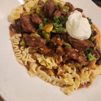 Biscotti's - Watered stroganoff - A dish of beef stroganoff features tender beef pieces, green onions, and a rich sauce served over cooked egg noodles. A dollop of sour cream is placed on top, garnished with chopped green onions. The presentation is on a white plate, highlighting the hearty and savory nature of the dish. - Cafes Near Me - Reno, Nevada
