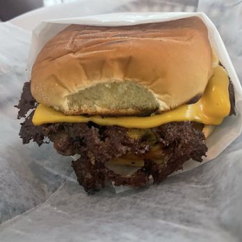 Mina Bistro - A cheeseburger with a soft, toasted bun, melted cheese, and crispy bacon strips. The burger is partially wrapped in white paper, with the bun slightly squished and the bacon visibly crispy and dark. The image focuses on the burger, showing the ingredients clearly, with a neutral background. - Venezuelan Near Me - Miami, Florida