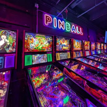 Free Play Bar Arcade - A series of pinball machines are arranged in a row inside a dimly lit arcade, each with vibrant, illuminated screens and colorful lighting effects. Above the machines, a neon sign spells out "PINBALL" in multicolored lights. The environment features dark walls and ceiling, emphasizing the bright, flashing lights of the arcade games, creating a lively and energetic atmosphere. - Bars Near Me - Providence, Rhode Island