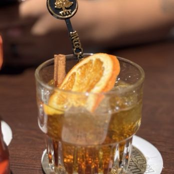 Buzzard Bar - Old fashioned - A clear glass filled with iced tea or a similar drink contains ice cubes, orange slices, and a cinnamon stick. The glass is placed on a round coaster on a wooden table. In the background, a person’s arm is visible, resting on the table, and part of a bottle can be seen on the left side of the image. - Bars Near Me - Ridgedale, Missouri