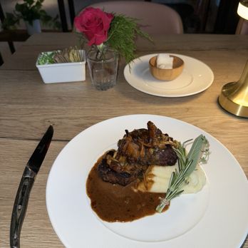Michael's - Petite Filet Diane - A dish consisting of a cooked steak topped with a mushroom sauce is presented on a white plate. The steak is accompanied by a portion of mashed potatoes, garnished with a sprig of rosemary. The setting includes a wooden table with a small vase of pink roses, a butter dish, and a lamp, indicating a restaurant dining environment. - Seafood Near Me - St. Augustine, Florida