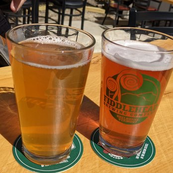 Station 101 - Beer.. yum - Two glasses of beer are on a wooden table, each on a green coaster. One glass contains a light-colored beer, and the other has a slightly darker beer with a logo for Fiddlehead Brewing Company. The background shows an outdoor seating area with black chairs, tables, and a lattice fence, suggesting a patio setting. - Beer Bar Near Me - Milford, New Hampshire