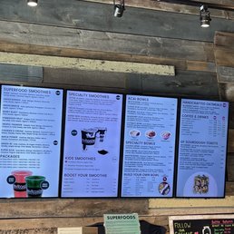 menus on the wall of a restaurant - Three digital menu screens display various food and drink options including smoothies, acai bowls, oatmeal, and toasts at Revitasize in Markham, ON. The menu includes categories such as superfood smoothies, kids smoothies, specialty bowls, and handcrafted oatmeal. Below the screens, there is a small counter with a paper menu titled 'Superfoods,' a black bag, and a small display stand. The background features a rustic wooden wall with track lighting overhead, creating a casual, modern atmosphere.