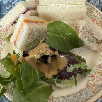 Lovey's Tea Shoppe - A porcelain plate with floral and gold detailing contains three finger sandwiches with different fillings, including ham, cheese, and a creamy mixture. A small side salad with mixed greens, dark leafy lettuce, and a drizzle of dressing is also on the plate. The plate is placed on a blue patterned tablecloth, and part of a matching teapot with a strawberry design is visible in the background. - British Near Me - Pacifica, California
