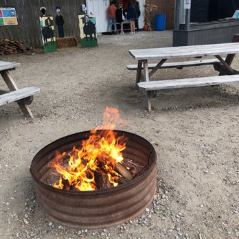 Schuster's Pumpkin Patch - The image shows a fire pit with a visible flame on a gravel ground in an outdoor setting. Surrounding the fire pit are wooden picnic tables, some with benches. In the background, there is a small snack shack with a sign, and several people are standing in line or near the shack. The area appears to be part of a fair, festival, or outdoor event space. - Pumpkin Patches Near Me - Elyria, Ohio