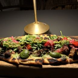 Jerk Lamb Flatbread - A flatbread with toppings including meat, green sauce, herbs, and vegetables, served on a wooden board with a small lamp in the background.