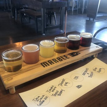 Marble Brewery - Six small glasses of beer, each with varying colors from pale yellow to dark brown, are arranged on a wooden flight paddle labeled 'MARBLE.' A menu below lists beer types including Double Cream Ale, Double IPA, Double TIPA, and others. The background shows a bar area with tables and chairs, indicating an indoor brewery setting. - Breweries Near Me - Albuquerque, New Mexico