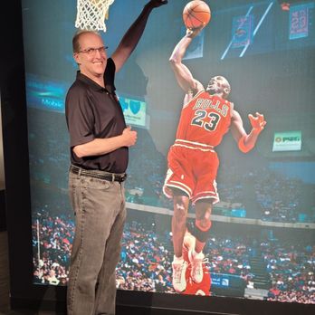 Skydeck Chicago - A man with glasses, wearing a black polo shirt and gray pants, stands indoors in front of a large digital display showing a basketball player from the Chicago Bulls mid-air, about to dunk. The display is part of the Skydeck Chicago attraction, which features panoramic city views and themed exhibits. - Observatories Near Me - Chicago, Illinois