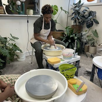 Seeing Green Studio - A woman is shaping clay on a pottery wheel in a room with various potted plants and decorative items. - Paint-Your-Own Pottery Near Me - Halethorpe, Maryland
