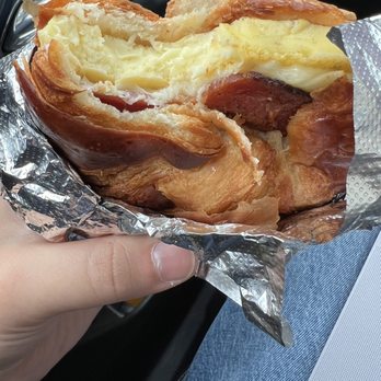 The Studio Cafe - Bacon, egg, and cheese on a croissant - A croissant sandwich containing bacon, egg, and cheese is shown partially unwrapped from aluminum foil. The sandwich is held in a person's hand, with the background showing the interior of a vehicle, including part of a dashboard and a person's lap in jeans. - Coffee & Tea Near Me - Woburn, Massachusetts
