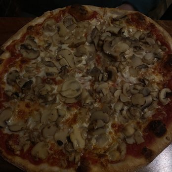 La Strada Restaurant - Mushroom pizza loaded with fresh mushrooms - A pizza topped with sliced mushrooms, melted cheese, and tomato sauce, with a golden-brown crust. The pizza is placed on a wooden table, and part of a person's arm is visible in the background. The image is taken indoors with low lighting. - Pizza Near Me - Millburn, New Jersey