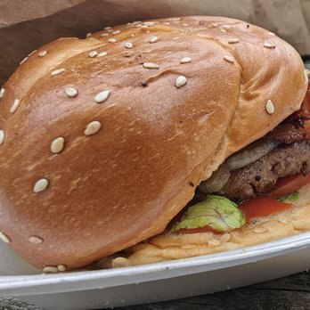 Paradise Pup - Burger - A burger featuring a sesame seed bun, beef patty, bacon, lettuce, and tomato is placed in a white takeout container. The burger appears freshly assembled with visible ingredients and is set on a wooden surface, with a brown paper bag in the background. - Fast Food Near Me - Des Plaines, Illinois