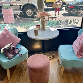 Hibiscus Brew - Inside a café, pastel blue chairs with pink pillows surround a marble table with a vase of pink flowers and drinks. Outside, a street scene with trucks, cars, and a person carrying a box is visible. - Cafes Near Me - Brooklyn, New York