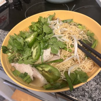 Việt kitchen - #16 - A bowl of Vietnamese pho features rice noodles, bean sprouts, cilantro, green onions, and slices of meat, garnished with fresh herbs and vegetables. The dish is placed on a kitchen countertop with a stove and other kitchen items visible in the background. Chopsticks rest on the bowl, ready for eating. - Vietnamese Near Me - Albuquerque, New Mexico