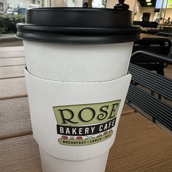Rose Bakery Cafe - A disposable coffee cup with a black lid and a white sleeve featuring the Rose Bakery Cafe logo, placed on a wooden table. - Bakeries Near Me - Orange, California