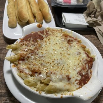 Fazoli's - Breadsticks and ziti with meat sauce. - A baked pasta dish featuring penne pasta with melted cheese and tomato sauce in a white ceramic baking dish. The dish is placed on a wooden table, with a side of breadsticks on a white rectangular plate behind it. Additional items include a plastic container, a receipt, and condiments, indicating a casual dining setting. - Italian Near Me - Amarillo, Texas