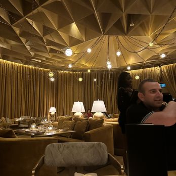 Ultra - Indoor dining restaurant - The image shows a restaurant interior with gold curtains lining the walls and plush beige sofas arranged for dining. The ceiling has a geometric, textured design with hanging spherical lights connected by chains. Several table lamps with white shades are placed on side tables, providing warm lighting. The tables are set with glassware, plates, and cutlery, indicating a prepared dining environment. There are a few patrons and staff visible, including a man and a woman engaged in conversation. - Salad Near Me - Toronto, Ontario