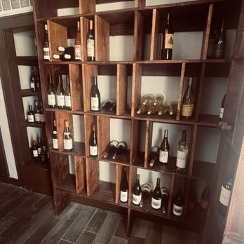 Pilon - A wooden wine rack with multiple compartments and shelves is filled with various wine bottles, some lying horizontally and others standing upright. The rack is positioned in a room corner with a dark wood floor and a white wall. Additional bottles are placed on top of the rack, and a small section of a door or cabinet is visible to the left. - Beer, Wine & Spirits Near Me - El Paso, Texas