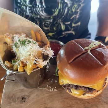The Kitchen Table - Kitchen Table Burger with Garlic Parmesan Fries - A cheeseburger with a beef patty, melted cheddar cheese, and a soft bun with a toothpick, placed on a wooden serving board. Next to it is a metal cup of fries topped with shredded cheese, chopped chives, and grated cheese, with a small container of ketchup behind. The background shows part of a person wearing a dark shirt and a watch. - American Near Me - Clermont, Kentucky
