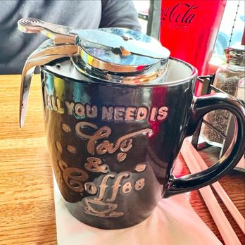 Market & Maple - A black ceramic coffee mug with embossed text reading 'All you need is love & coffee' and a small cup illustration, placed on a wooden table. The mug has a metal lid with a spoon resting on top. In the background, there is a red Coca-Cola cup, a glass jar, and a person wearing a gray shirt. The setting appears to be a casual dining environment. - Breakfast & Brunch Near Me - Dyer, Indiana