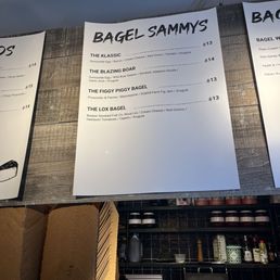 Bagel options - A printed menu board at Bagel Sammys in Boston, MA, lists four bagel options: The Klassic, The Blazing Boar, The Figgy Piggy Bagel, and The Lox Bagel. Each option includes ingredients such as eggs, meats, cheeses, and toppings, with prices ranging from $13 to $15. The background shows a wooden panel and part of the shop interior with shelves and kitchen items. A small sign at the bottom right requests notifying servers of allergies before ordering.