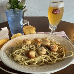 Scallops Linguine - Linguine with scallops and garlic bread are served on a disposable plate, accompanied by a Stella Artois beer and a glass of water on a wooden table.