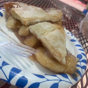 Java Junction - Apple pie - A disposable paper plate holds slices of apple pie with a golden, flaky crust and apple filling, topped with a light dusting of powdered sugar. The pie is served outdoors on a metal table, with a plastic fork resting on the plate. The background shows a red and white checkered tablecloth and outdoor seating, indicating a casual dining setting. - Bed & Breakfast Near Me - Madrid, New Mexico