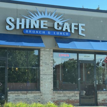 Shine Cafe - The image shows the exterior of Shine Cafe in Troy, MI, with a prominent sign displaying the cafe's name and a sunburst logo. Blue awnings extend over the windows and entrance. The glass door has an 'Open' sign illuminated in pink, and the building's facade includes brick and stucco elements. Green plants are visible at the base of the storefront, and the sky is clear and blue. - Breakfast & Brunch Near Me - Troy, Michigan