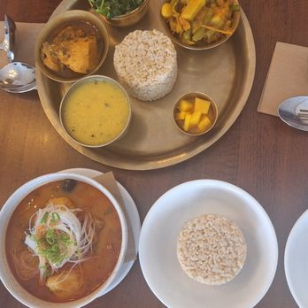 Momo Valley - dal bhat, phing sha - The image shows a Himalayan restaurant meal with a main tray containing a round serving of rice, vegetable curries, and a bowl of yellow soup. A small bowl of mango chunks is also present. Additionally, there is a bowl of noodle soup with green onions on top, placed on a wooden table with utensils and a glass of water nearby. - Himalayan/Nepalese Near Me - Beacon, New York