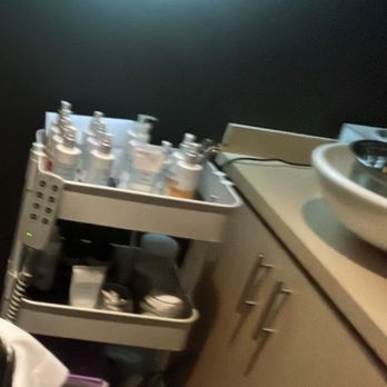 Pure + Simple - A mobile cart with multiple bottles and containers is situated next to a sink and a cabinet with metallic handles. The room has dark-colored walls and appears to be a professional or clinical setting, possibly a spa or medical room. - Day Spas Near Me - Toronto, Ontario