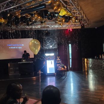 PlugIN Karaoke - DJ prepping - An indoor event space features a stage with balloons hanging from a truss, a large screen, and people seated at tables, possibly for a celebration. - Karaoke Near Me - Hallandale Beach, Florida