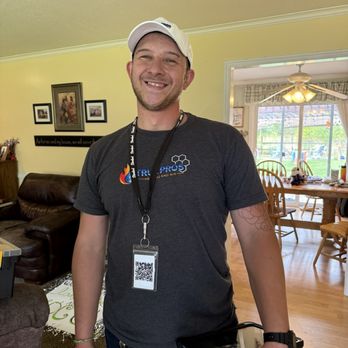 True Pros - A man with a beard and short hair, wearing a white cap, a black True Pros T-shirt, and a lanyard with an ID badge, stands indoors in a living room. He is smiling and has a tattoo on his left arm. The background shows a dining area with a wooden table and chairs, a ceiling fan, and large windows with curtains, revealing an outdoor patio area. - Heating & Air Conditioning/HVAC Near Me - Layton, Utah