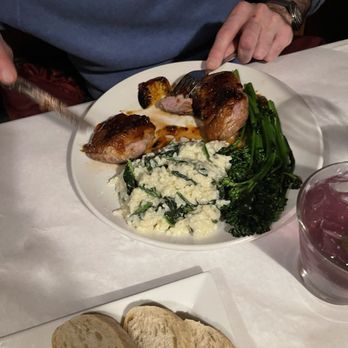 Glenwillow Grille - Duck - A dinner plate contains two pieces of cooked meat, green vegetables, and mashed potatoes with spinach. A person is cutting the meat, and there is bread on a separate plate. - American Near Me - Solon, Ohio