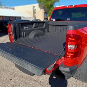 Auto Depot - My Rhino lined truck bed. - The image depicts the open cargo area of a red pickup truck with a black bed liner, parked outdoors on a paved surface. The truck bed is empty and features a textured surface for durability. The background includes a building, some parked cars, a tree, and a clear blue sky, indicating a sunny day. The photo is focused on the truck bed, with the tailgate lowered, providing a clear view of the interior space. - Auto Detailing Near Me - Albuquerque, New Mexico
