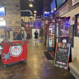 Entry to place. - The image shows the interior of Backporch Drafthouse, a sports bar and restaurant, decorated with Christmas ornaments including a Christmas tree and garlands. Multiple TV screens display sports events. The space features a stone wall, string lights, and a sign indicating the bar's name. A staff member is at the entrance, and a woman is walking further into the establishment. The decor includes festive elements and seating areas for patrons.