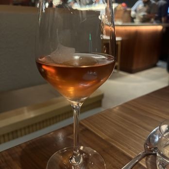 Uchi Miami - Rosé - A glass of rosé wine is placed on a wooden table in a restaurant, with a blurred background showing other patrons and a bar area. - Japanese Near Me - Miami, Florida
