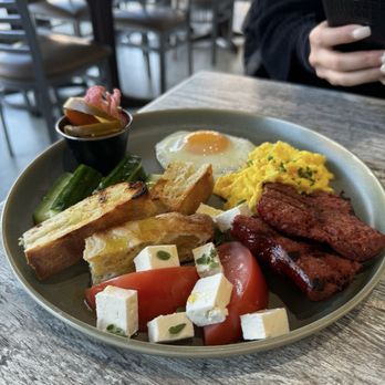 Marson Caffè - Balkan Breakfast - A breakfast plate featuring a fried egg, scrambled eggs, grilled meat, toast, cucumber, tomato, and feta cheese, with a small side of pickled vegetables. - Cafes Near Me - Brooklyn, New York