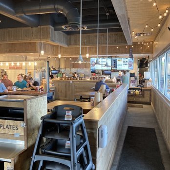 City Barbeque - The image shows the interior of a casual dining restaurant with a modern rustic design. Wooden paneling covers the walls and ceiling, complemented by hanging light bulbs and exposed ductwork. Digital menu screens are visible behind the counter, where staff are preparing orders. Customers are seated at tables, some engaged in eating or conversation. A high ceiling and large windows on the right side provide natural light, creating an inviting atmosphere. - Caterers Near Me - Kettering, Ohio