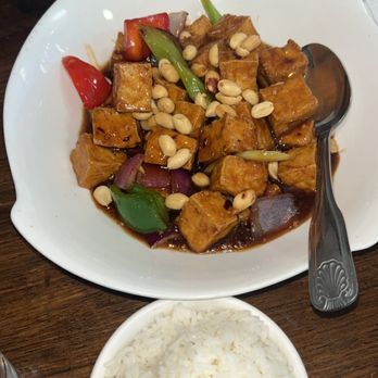 Veggie House - Kung Pao Tofu - A white bowl contains tofu cubes stir-fried with red and green bell peppers, onions, and peanuts, coated in a dark sauce. A separate small bowl holds steamed white rice. The setting appears to be a restaurant table with a spoon placed on the plate and a person's hands visible in the background. - Vegan Near Me - Chicago, Illinois