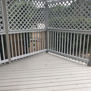 Evdecks Construction - Replaced lattice, pergola and stained - The image shows a corner section of a wooden deck painted in light color, with vertical white railing posts and lattice panels on the sides and back. A pergola with slatted roof is installed above, providing partial shade. A wooden fence and greenery are visible beyond the deck, indicating an outdoor residential area. - Fences & Gates Near Me