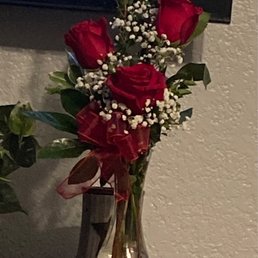 The second, corrected bouquet. - A bouquet of three red roses accompanied by sprigs of baby's breath is arranged in a clear, slightly curved glass vase. The vase is positioned on a wooden surface against a textured wall, with a television screen visible in the background. The arrangement includes green foliage and a red ribbon tied around the vase, creating a simple yet elegant display.