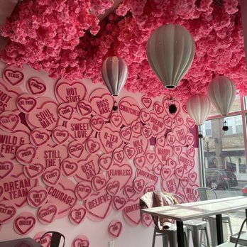 Bober Tea - The image shows a cafe interior with a ceiling covered in dense pink floral arrangements. The wall is decorated with numerous pink heart-shaped signs displaying various words and phrases. Several white balloon-shaped pendant lights hang from the ceiling. Large windows on the right side allow natural light to illuminate the space, revealing a view of parked cars and buildings outside. The seating includes a high table with chairs and a cushioned chair near the window. - Donuts Near Me - Carmel, Indiana
