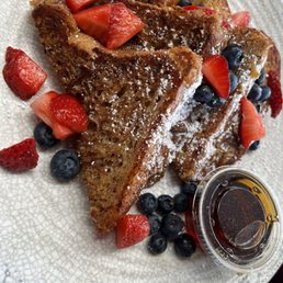 Fruit loaded French toast - A plate of French toast slices is presented with toppings including sliced strawberries, blueberries, and a dusting of powdered sugar. A small container of syrup is placed on the side. The dish is served on a white textured plate, highlighting the golden-brown color of the French toast and the vibrant colors of the fruit.